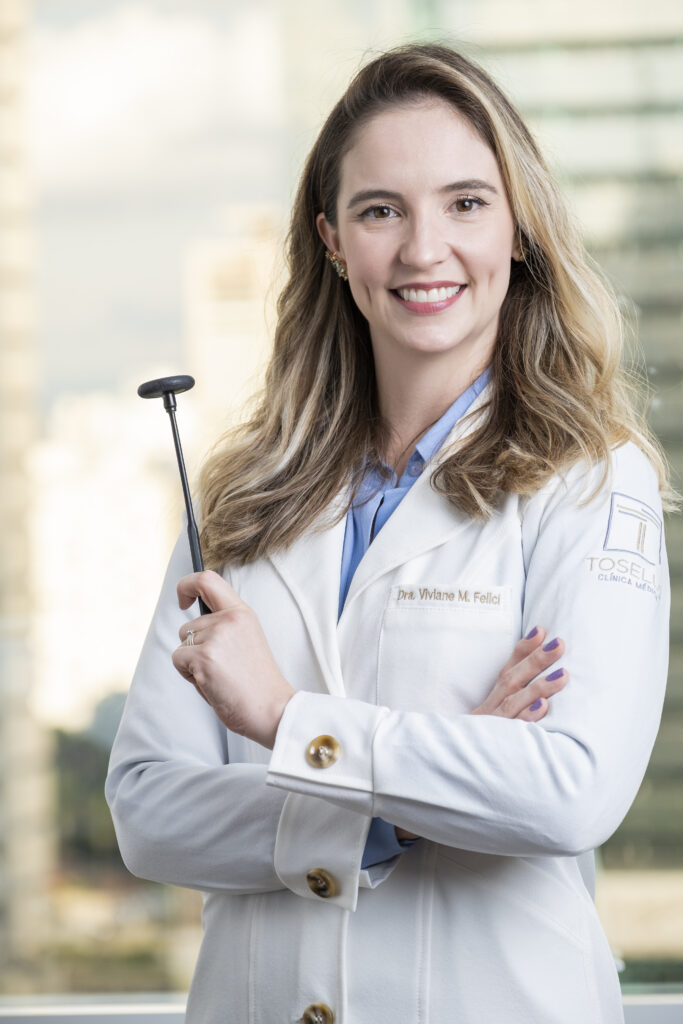 Foto da Dra Viviane, neurologista, de jaleco, segurando um martelo neurológico em seu consultorio