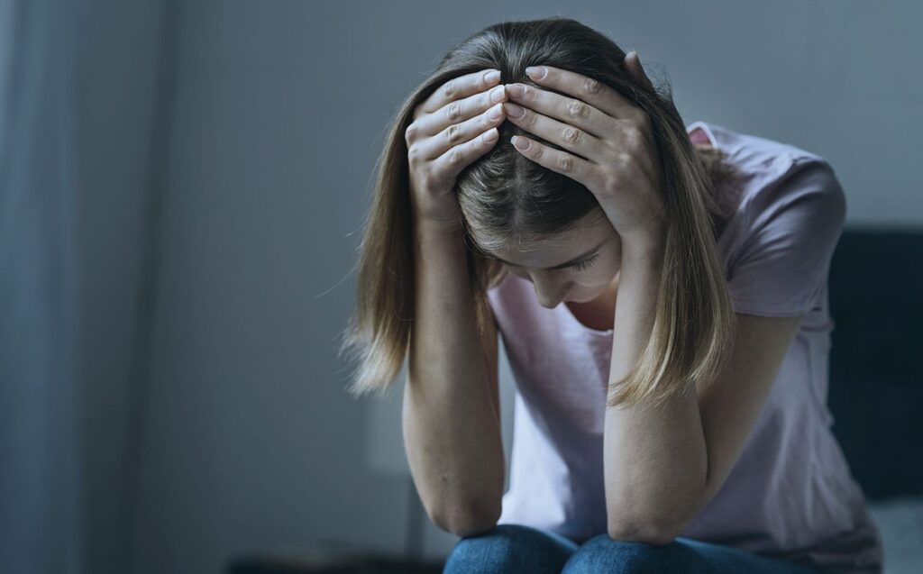 Mulher sentada com as mãos na cabeça sentindo dor de cabeça em um quarto na penumbra
