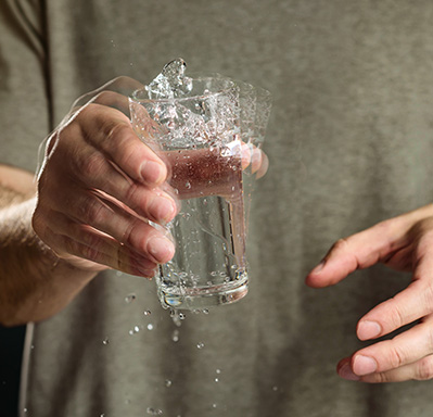 Homem com tremor nas mãos, segurando um copo de vidro com água que está balançando e derrubando água