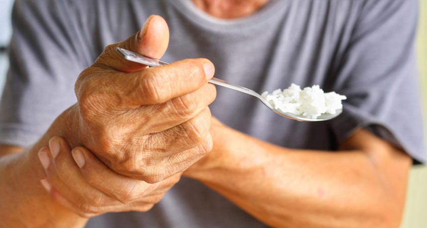 Homem com tremor nas mãos segurando uma colher cheia de arroz, ele apoia uma mão com a outra para diminuir o tremor