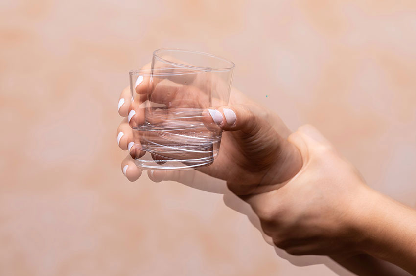 Mulher com tremor nas mãos, segurando um copo de vidro com água que está balançando
